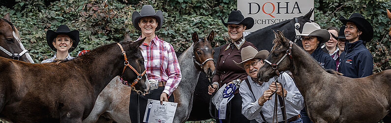 Mehrere Personen mit Westernhüten stehen mit Pferden vor einem grünen Hintergrund; einige halten Schleifen und Urkunden. Im Hintergrund ein Schild mit der Aufschrift „Höveler DOHA Zuchtschau“.