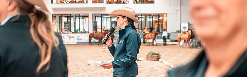 Eine Sprecherin mit Cowboyhut hält ein Mikrofon und spricht in einer Reithalle, während im Hintergrund Pferde und Zuschauer zu sehen sind. Die Szene zeigt eine Moderation oder Siegerehrung im Rahmen einer Pferdesportveranstaltung.