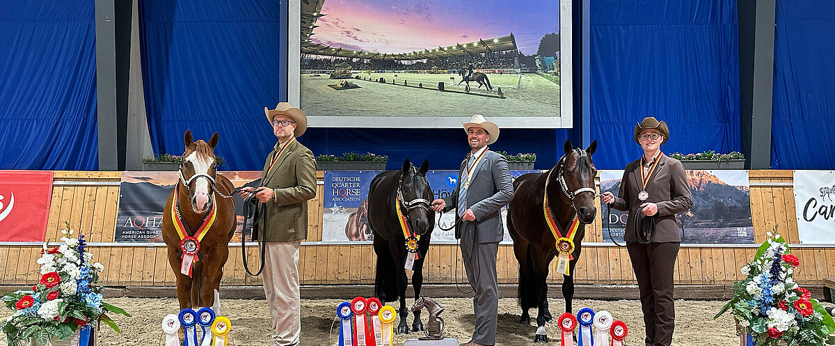 Drei Personen stehen mit drei Pferden bei einer Siegerehrung auf einem roten Teppich. Text: CONGRATULATIONS, 03-12.10.2024 AACHEN GERMANY, Q24, DQHA DEUTSCHE QUARTER HORSE ASSOCIATION.