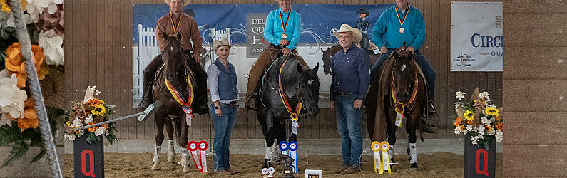 Siegerehrung in einer Reithalle: Drei Reiter sitzen auf ihren Pferden und tragen Medaillen um den Hals, flankiert von zwei weiteren Personen am Boden. Vor den Pferden liegen farbige Schleifen und Pokale auf einem roten Teppich. Links und rechts stehen dekorative Blumengestecke mit schwarzen Sockeln und rotem „Q“-Logo. Im Hintergrund sind Banner der Deutschen Quarter Horse Association und weiterer Sponsoren an der Hallenwand angebracht.